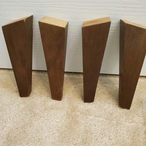 May include: Four wooden furniture legs, each with a trapezoid shape. The legs are a medium brown colour and have a flat top. They are arranged in a row on a light-coloured carpet, with a white wall in the background.