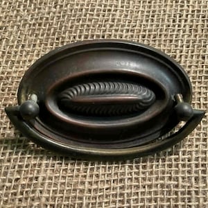 Puede incluir: Un tirador de cajón antiguo en tono bronce con una placa trasera ovalada y un diseño central texturizado. El tirador presenta un asa curva y dos pequeños pomos redondos a cada lado. El herraje está diseñado para muebles.