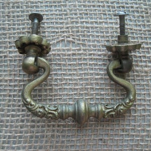 May include: Two antique brass drawer pulls with ornate, curved handles and decorative screw heads. The pulls have a vintage aesthetic, with a slightly tarnished finish, suggesting age and use. These are suitable for furniture restoration.
