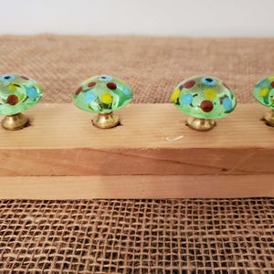 May include: Four mushroom-shaped cabinet knobs. Each knob has a green glass cap with colorful polka dots in blue, yellow, and red. The knobs are mounted on a light-colored wooden board with brass bases. The background is a textured, tan fabric.