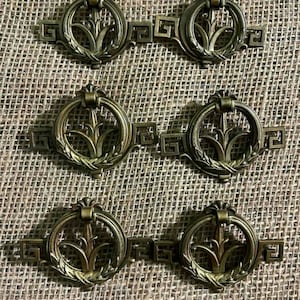 May include: Six antique bronze drawer pulls. Each pull features a circular handle with a central floral design and a rectangular base with a geometric pattern. The pulls are arranged on a textured, tan surface.