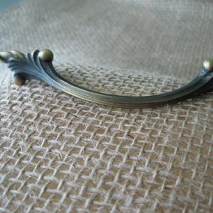 May include: An antique-style bronze cabinet pull handle. The curved handle features decorative leaf and ball accents on each end. The hardware is set against a textured, neutral-toned fabric background.