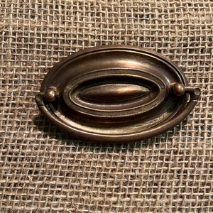 May include: An oval-shaped bronze drawer pull featuring a central raised horizontal bar surrounded by a textured ring and a smooth outer border. The hardware includes a hinged drop handle attached to the sides, showcasing a dark metallic finish.