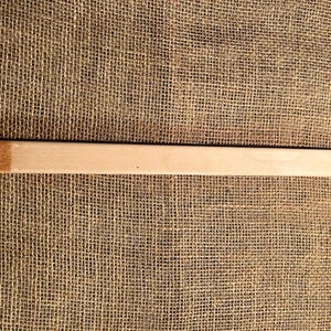 May include: A light-colored wooden bar with two drilled holes, one at each end. The bar has a rectangular shape and a smooth surface. The wood has a natural grain and a slight color variation at one end. The bar is set against a burlap background.