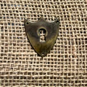 May include: A shield-shaped, antique brass keyhole cover with a keyhole design. The cover has a decorative top and three small holes for mounting. The keyhole cover is set against a textured, tan burlap background.