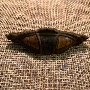 May include: A metal drawer pull with a dark, weathered bronze finish. The hardware features a central rectangular panel flanked by two triangular sections, all displaying a wood-grain pattern. The edges are decorated with a raised, scalloped border.