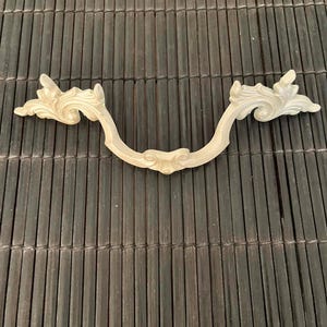 Pode incluir: Puxador de gaveta ornamentado, branco sujo, com um design curvo. A pega apresenta detalhes florais decorativos e rodopiantes em cada extremidade e um adorno central. Ferragens de material claro.