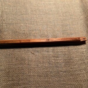 May include: A long, rectangular, wooden object with a dark brown finish. The object has a series of grooves and a small hole at one end. It is resting on a textured, tan fabric.