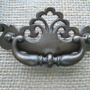 May include: Antique bronze-toned drawer pull with an ornate, symmetrical design. The pull features a curved handle and decorative flourishes, with mounting hardware on each end. This hardware is suitable for cabinets and furniture.