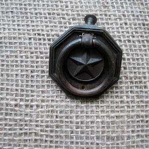 May include: Octagonal metal cabinet pull with a star-shaped design and a ring handle. The pull is a dark brown color and has a rustic finish.