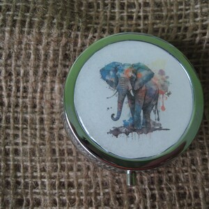 May include: A silver round trinket box with a colorful watercolor elephant design. The elephant is blue, green, orange, and yellow.