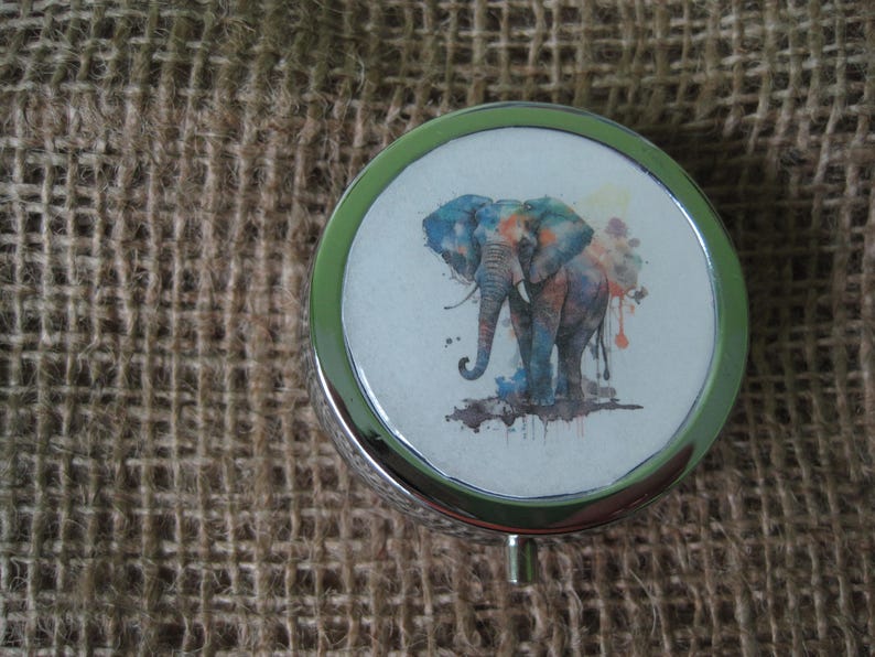May include: A silver round trinket box with a colorful watercolor elephant design. The elephant is blue, green, orange, and yellow.