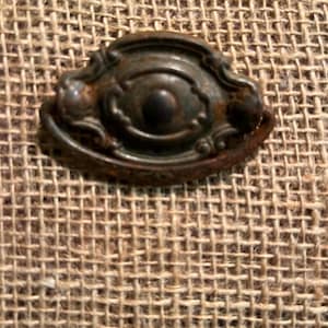 May include: A dark, weathered metal drawer pull featuring an oval-shaped backplate with ornate, raised scrollwork detailing and a central circular knob. A curved, semi-circular drop handle hangs from the base of the decorative mounting plate.