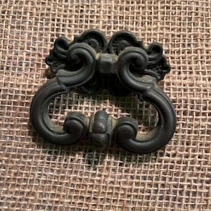 Könnte beinhalten: Ein dunkel bronzefarbener Schubladengriff aus Metall mit einem verzierten, symmetrischen Design, geschwungenen Kurven und einem zentralen Dekorelement. Der Beschlag hat eine verwitterte Oberfläche und einen beweglichen Griff.