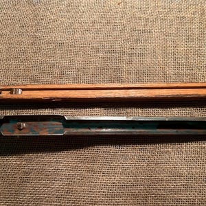 May include: Two long, narrow, rectangular components. The top piece is made of light brown wood with a visible grain and a small metal insert. The bottom piece is a dark, weathered metal channel with patches of blue paint and visible oxidation.