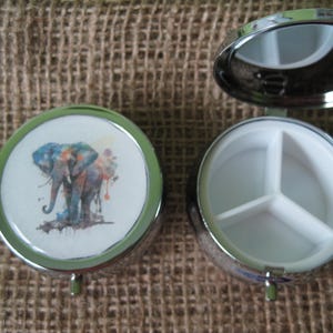 May include: A silver pill box with a watercolor elephant design on the lid and a three-compartment interior.