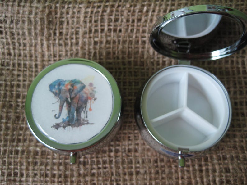 May include: A silver pill box with a watercolor elephant design on the lid and a three-compartment interior.