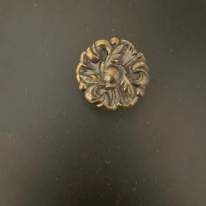 May include: A detailed, antique-style bronze-colored cabinet knob. The knob features an ornate floral design with intricate leaf and swirl patterns. The hardware is circular and appears to be made of metal.