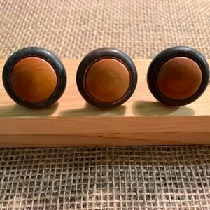Puede incluir: Tres pomos circulares con un inserto central de madera de color marr&oacute;n claro liso, rodeado por un anillo exterior de color negro mate oscuro. Estas piezas de herraje est&aacute;n dispuestas en una fila horizontal, mostrando un dise&ntilde;o bicolor.
