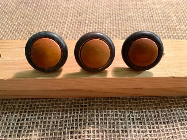 Puede incluir: Tres pomos circulares con un inserto central de madera de color marr&oacute;n claro liso, rodeado por un anillo exterior de color negro mate oscuro. Estas piezas de herraje est&aacute;n dispuestas en una fila horizontal, mostrando un dise&ntilde;o bicolor.
