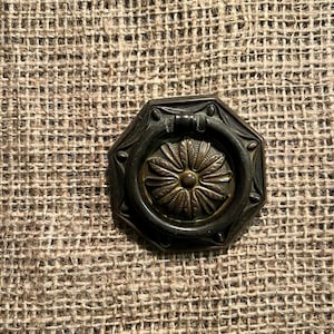 May include: An antique bronze-toned drawer pull with an octagonal frame and a central floral design. The pull features a ring handle and is set against a textured, tan burlap background. This hardware piece is suitable for furniture restoration.