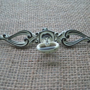 May include: An ornate, off-white and black drawer pull with a decorative design. The pull features a central ring handle and curved, scroll-like arms extending outwards. The hardware is likely for furniture or cabinetry.