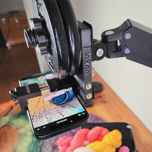 May include: A black phone is mounted on a tripod with a weight attached to the arm. The phone is recording a video of a colourful crocheted blanket.