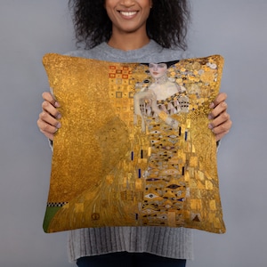 May include: A gold throw pillow with a painting of a woman in a gold dress with a geometric pattern. The painting is in the style of Gustav Klimt.