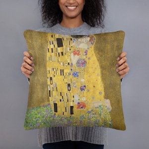May include: A square throw pillow with a gold background featuring a painting of two figures embracing. The painting is in a style reminiscent of Gustav Klimt's "The Kiss" with a mosaic-like pattern of gold, black, and white squares. The figures are wearing robes with colorful floral patterns.