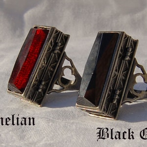 Könnte beinhalten: Zwei silberne Ringe mit verzierten Designs. Der Ring links hat einen roten Karneol-Stein. Der Ring rechts hat einen schwarzen Onyx-Stein. Die Ringe sind mit "Karneol" und "Schwarzer Onyx" beschriftet.