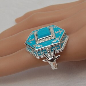 May include: A silver ring with a turquoise blue enamel box design. The box is decorated with silver accents and has a small animal figure on the side.