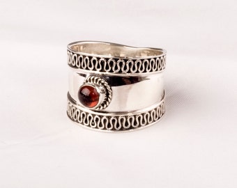 May include: A silver ring with a red gemstone set in a decorative bezel. The ring has a wide band with a textured design.