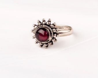 Può includere: Un anello d'argento con una pietra preziosa rossa incastonata in un design a forma di sole. L'anello è contrassegnato "925" sulla fascia.