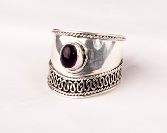 May include: A wide silver ring with an oval, deep purple gemstone set in the center. The ring has a decorative, wavy pattern along the bottom edge and a twisted rope design along the top and bottom edges.