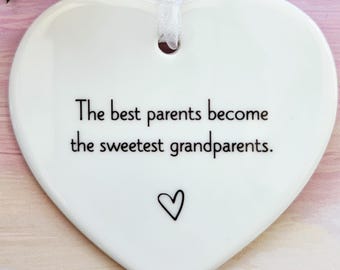 May include: White heart-shaped ceramic ornament with the text "The best parents become the sweetest grandparents." A small heart is drawn below the text. A white ribbon is attached for hanging. An orange rose and green leaves are in the background.