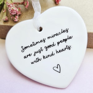 Può includere: Ornamento in ceramica bianca a forma di cuore con la scritta "Sometimes miracles are just good people with kind hearts". Un piccolo cuore nero è disegnato sotto il testo. Un nastro bianco è attaccato per appenderlo. Fiori rosa sullo sfondo.