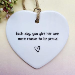 May include: White heart-shaped ceramic ornament with a jute string. The ornament has the text "Each day, you give her one more reason to be proud." and a small heart symbol. Decorative flowers are in the background.