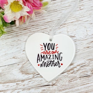 May include: White ceramic heart-shaped ornament with a white ribbon. The ornament has black text that reads "YOU are an AMAZING mother" with red hearts on either side of the word "mother".