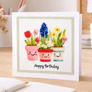 May include: A white birthday card featuring a watercolour illustration of three flower pots. The pots are pink, red, and blue, each with a smiling face and different flowers. The text "Happy Birthday" is below.