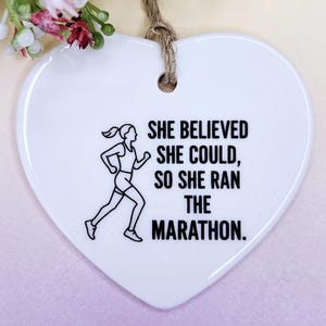May include: White heart-shaped ornament with a running figure graphic and the text "SHE BELIEVED SHE COULD, SO SHE RAN THE MARATHON." The ornament has a jute string for hanging. Decorative floral accents are visible at the top.