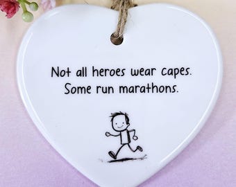 Puede incluir: Adorno de cerámica blanco en forma de corazón con el texto "No todos los héroes llevan capa. Algunos corren maratones". Debajo del texto hay una pequeña caricatura en blanco y negro de una persona corriendo.