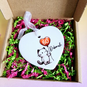 May include: A white heart-shaped ornament with a cartoon cat holding a red balloon with the number 60. The name "Deborah" is written on the ornament. The ornament is in a cardboard box with pink and green paper filler.