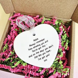 May include: A white heart-shaped ornament with a ribbon, inside a cardboard box filled with pink and green paper shreds. The ornament has the text "Abbie, Always remember how amazing you are! You've got this - and on the days you don't feel like you have... I've got you!"