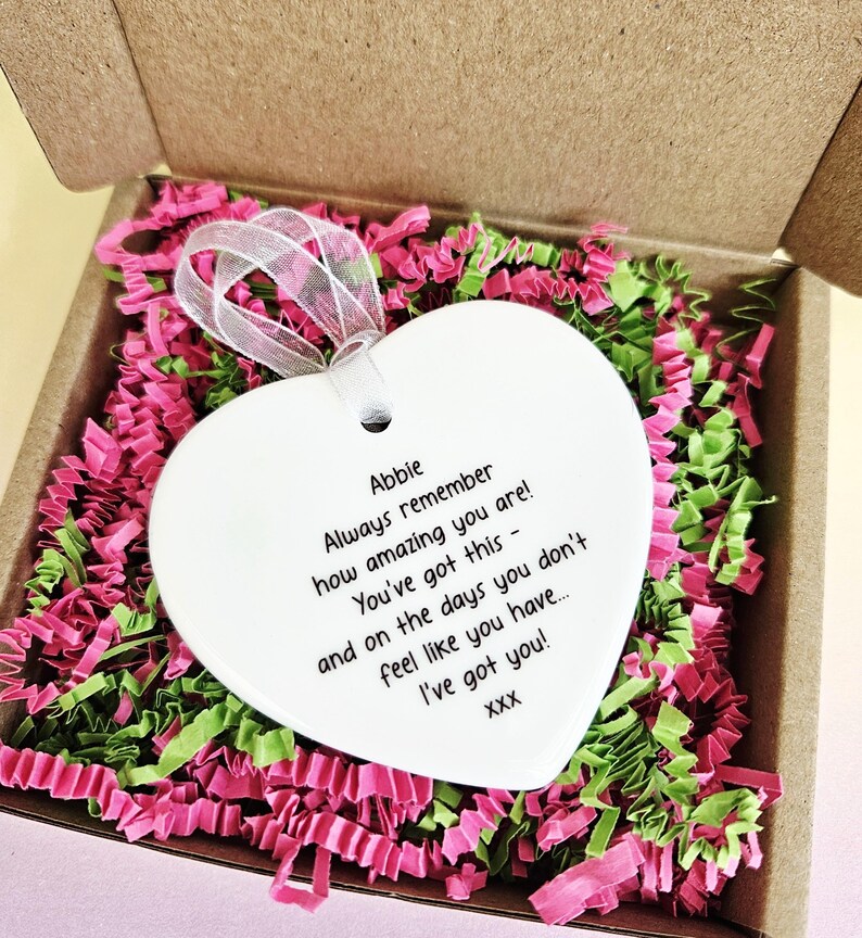 May include: A white heart-shaped ornament with a ribbon, inside a cardboard box filled with pink and green paper shreds. The ornament has the text "Abbie, Always remember how amazing you are! You've got this - and on the days you don't feel like you have... I've got you!"