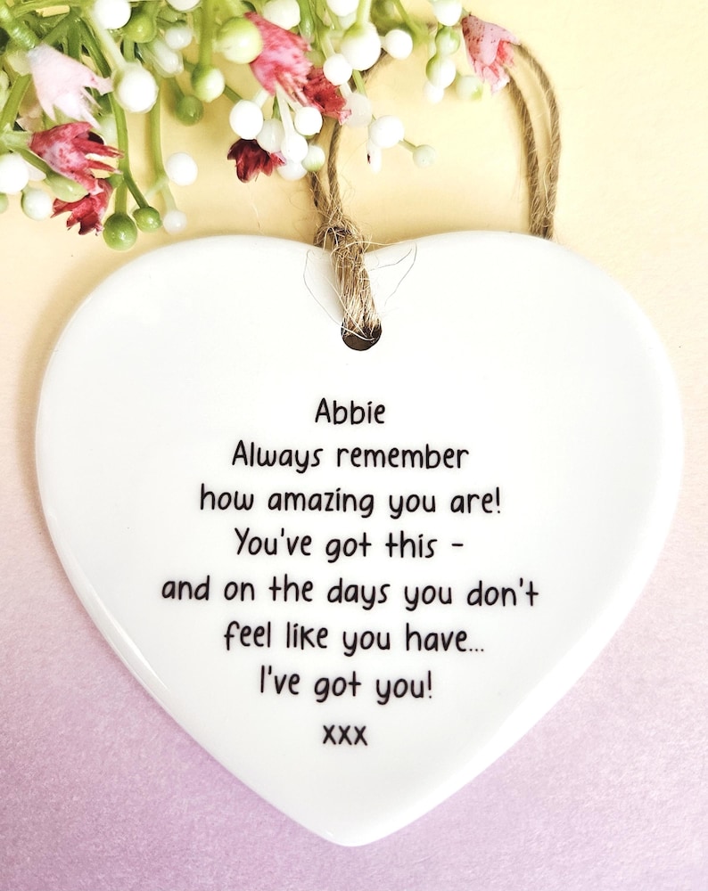 May include: A white heart-shaped ceramic plaque with a jute string for hanging. The plaque features the name "Abbie" and an inspirational message printed in black. Floral accents with white and red flowers are at the top.