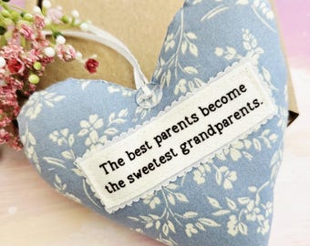 May include: A heart-shaped, light blue fabric ornament with a white floral pattern. A white rectangular patch with the text "The best parents become the sweetest grandparents" is sewn on the front. A white ribbon is attached for hanging.