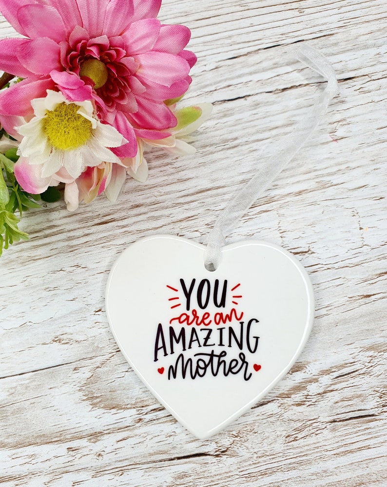 May include: White ceramic heart-shaped ornament with a white ribbon. The ornament has black text that reads "YOU are an AMAZING mother" with red hearts on either side of the word "mother".