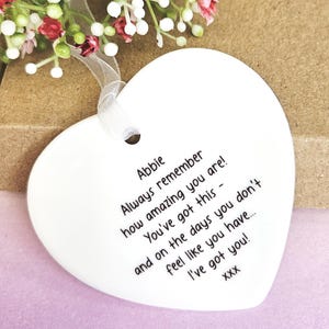 May include: A white heart-shaped ceramic ornament with a ribbon for hanging. The ornament has black text that reads "Abbie Always remember how amazing you are! You've got this - and on the days you don't feel like you have... I've got you!"