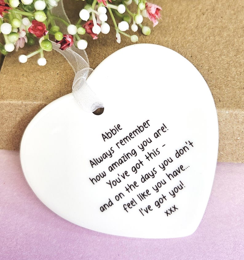 May include: A white heart-shaped ceramic ornament with a ribbon for hanging. The ornament has black text that reads "Abbie Always remember how amazing you are! You've got this - and on the days you don't feel like you have... I've got you!"