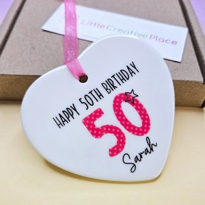 May include: White ceramic heart-shaped ornament with the text "Happy 50th Birthday" and the number "50" in pink with white polka dots. The name "Sarah" is written below the number.